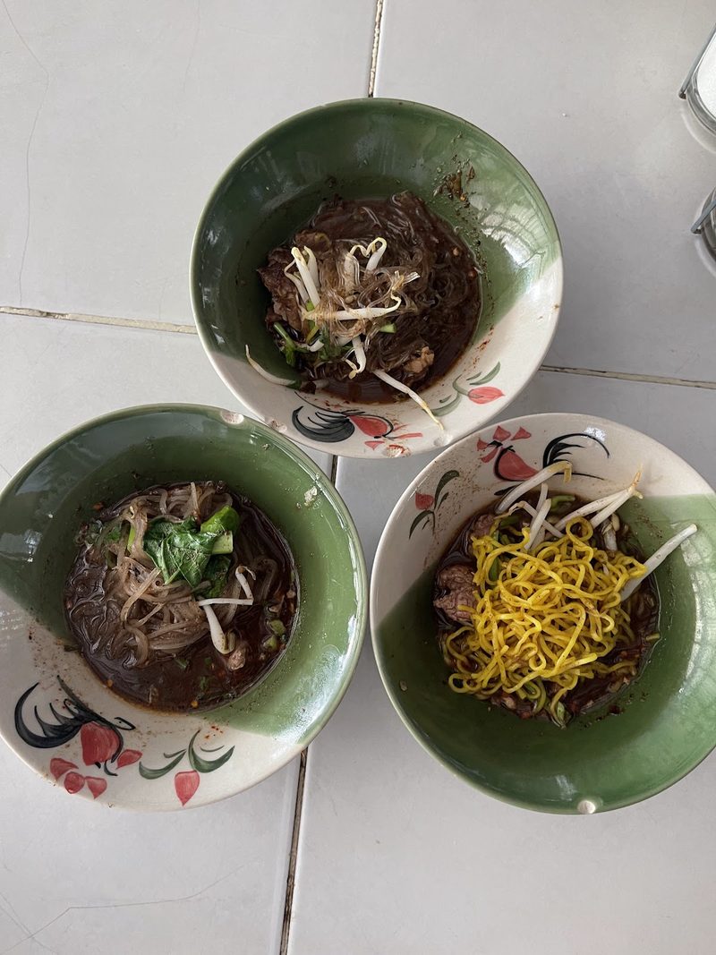 ก๋วยเตี๋ยวเรือสามชามน้ำซุปสีเข้ม มีเนื้อ ลูกชิ้น และผักบุ้ง