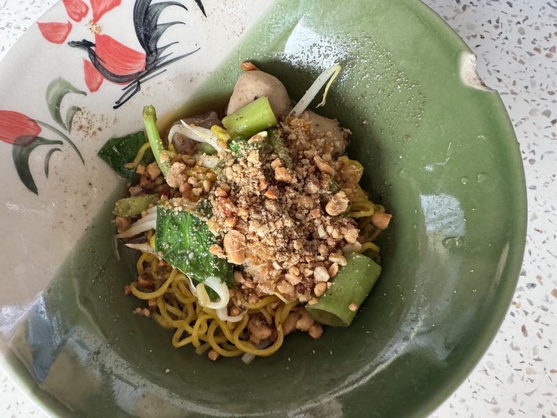 บะหมี่แห้งในชามเขียว โรยถั่วป่น พริก มีลูกชิ้นและผักบุ้ง