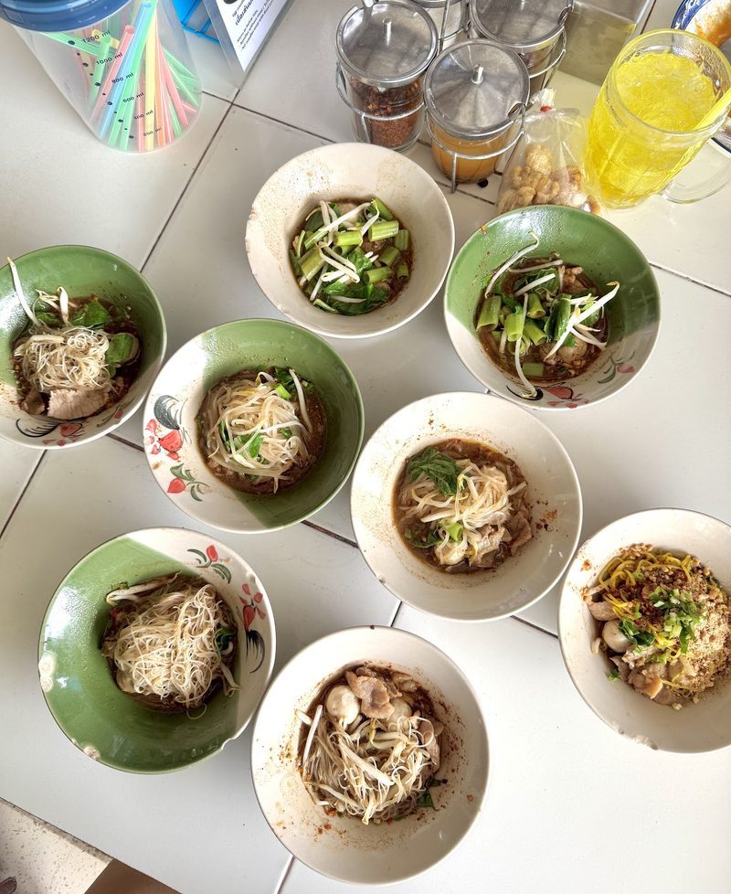 ก๋วยเตี๋ยวเรือชามเล็กเต็มโต๊ะ กับเครื่องปรุงและเครื่องดื่มเย็น