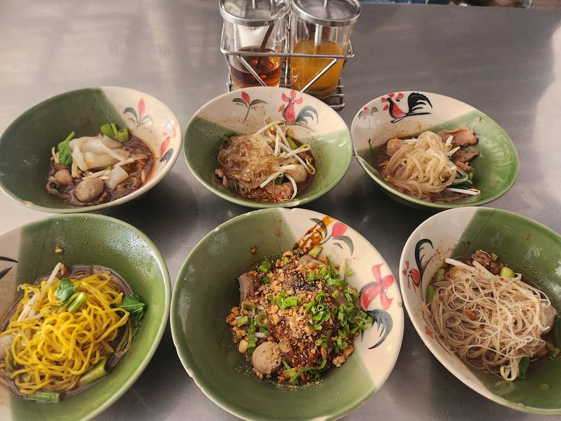 ก๋วยเตี๋ยวเรือชามเล็กหกชามบนโต๊ะสเตนเลส มีทั้งเส้นเล็ก วุ้นเส้น บะหมี่ และชามแห้งโรยถั่ว